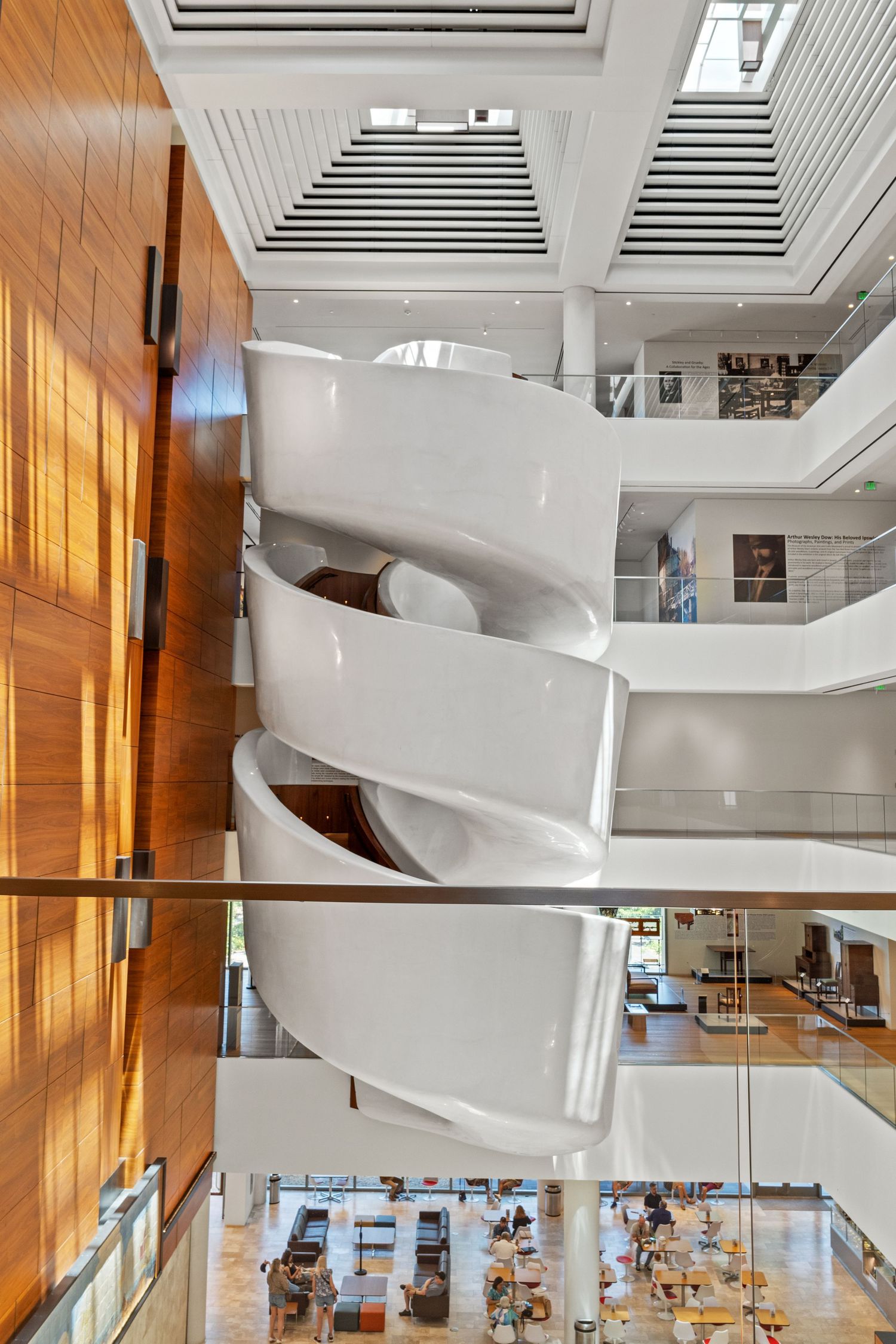 Modern museum interior featuring sleek white spiral staircase and warm wood paneling with natural lighting.