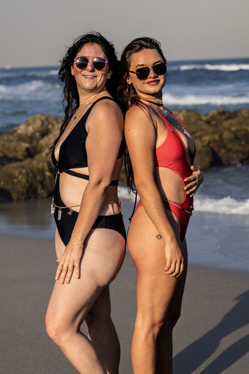 Two women in swimsuits pose together on beach with ocean and rocks in background.