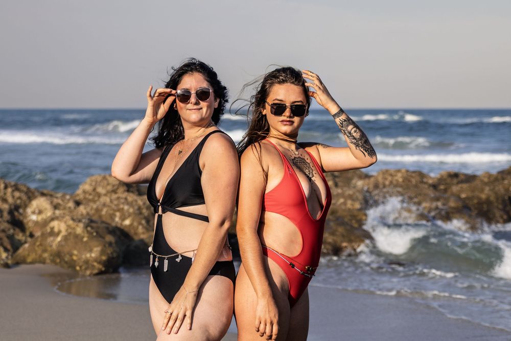 Two friends in swimwear pose playfully on a rocky beach with ocean waves in the background.