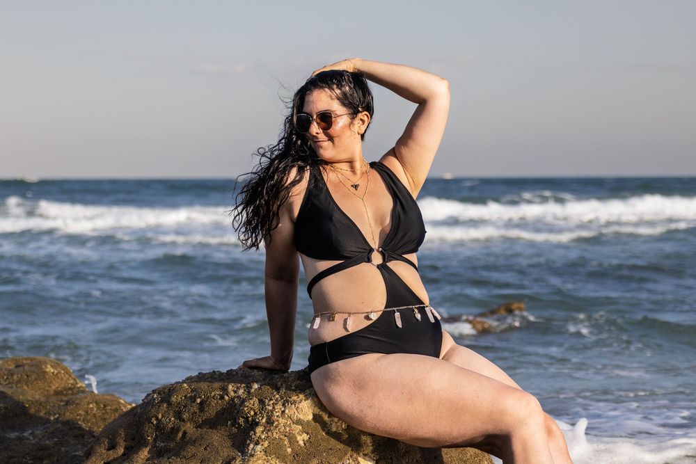 Woman in dark swimwear poses against ocean backdrop on rocky beach.