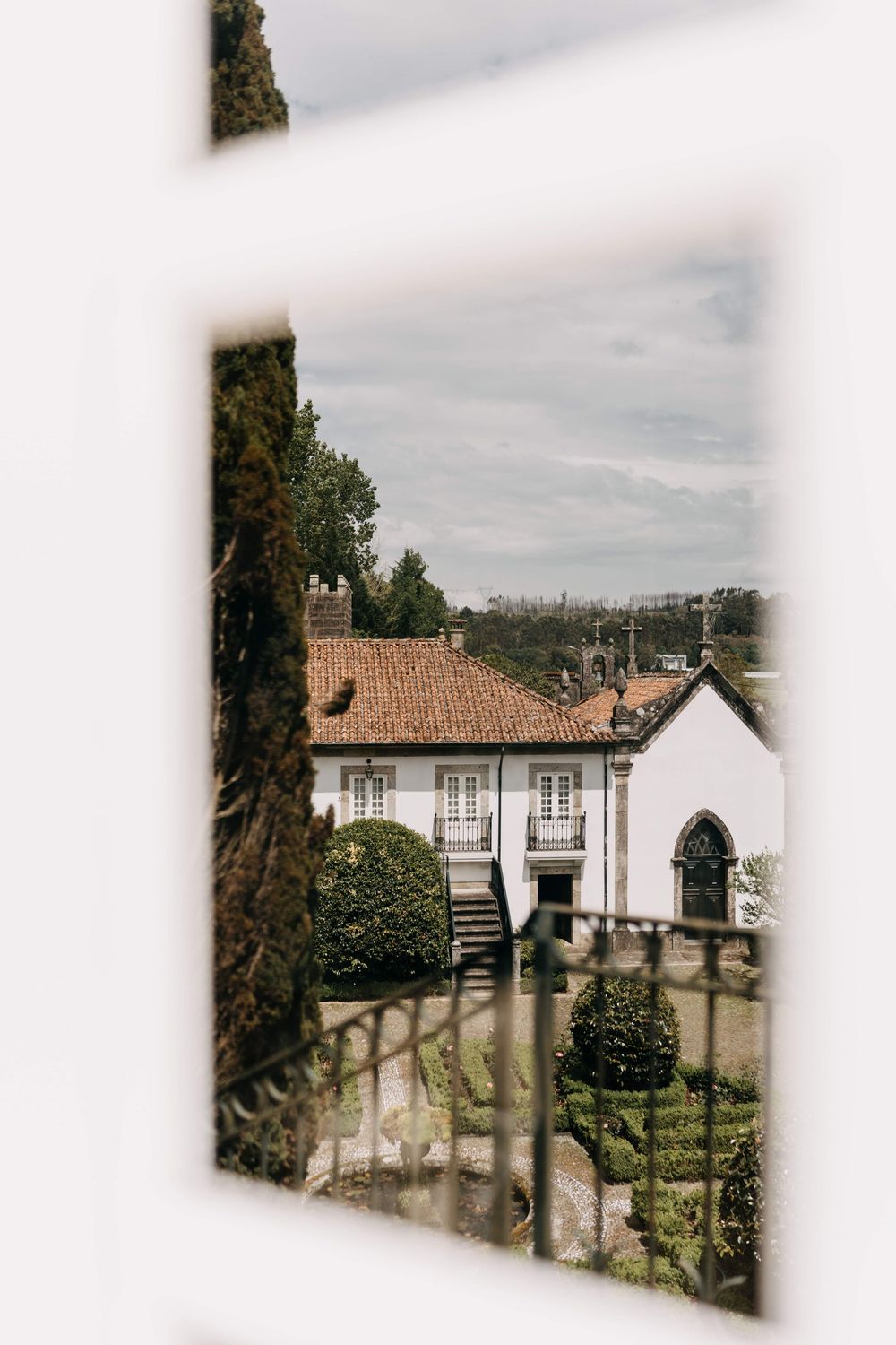 View through window frame of traditional Portuguese house with terracotta roof and small chapel in garden setting.