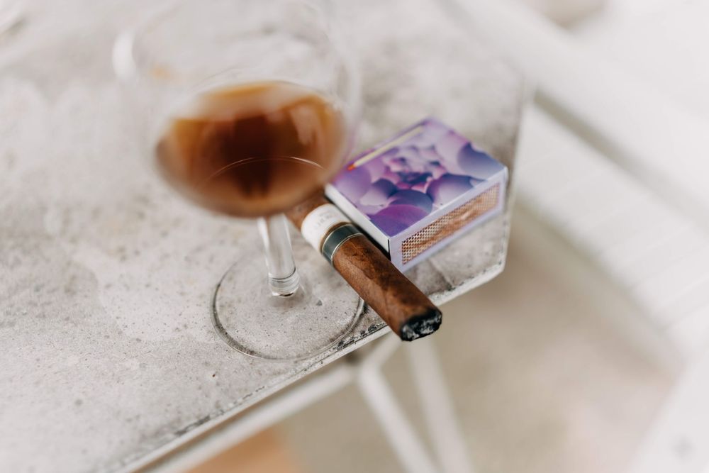 A cigar rests beside a wine glass and perfume bottle on a white textured surface.