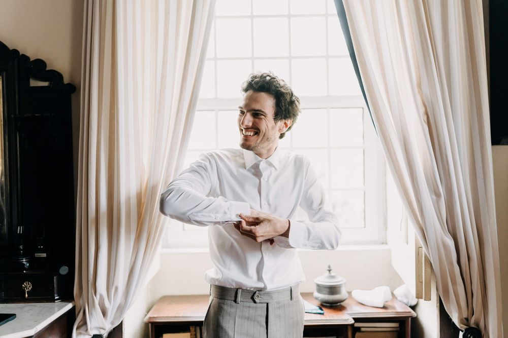 A person in a white dress shirt checks their watch while standing by a window with white curtains.