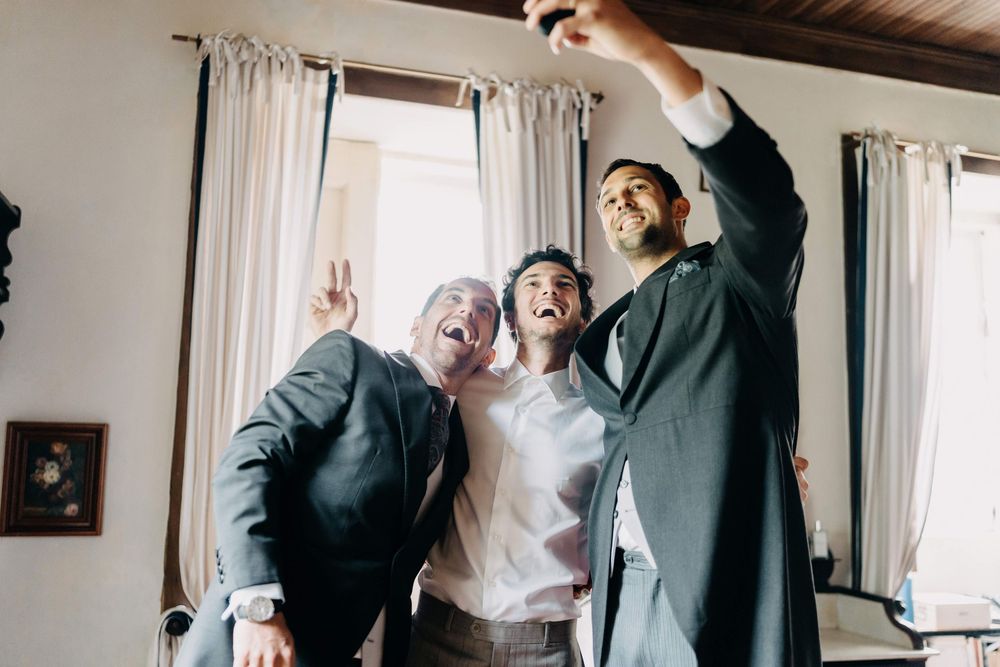 Three people in formal attire share a joyful moment striking celebratory poses inside a room with white curtains.