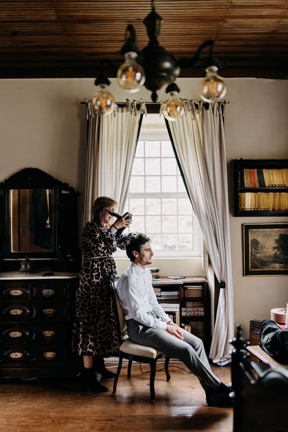 A vintage-themed room with elegant curtains and chandelier as someone styles the other's hair before a large window.