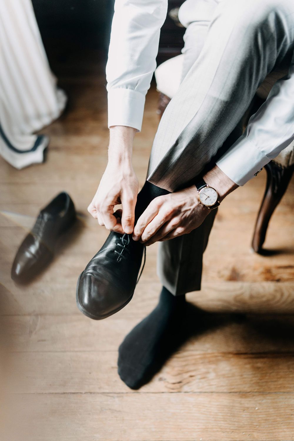 Person in formal attire tying black leather dress shoes while sitting on wooden floor.