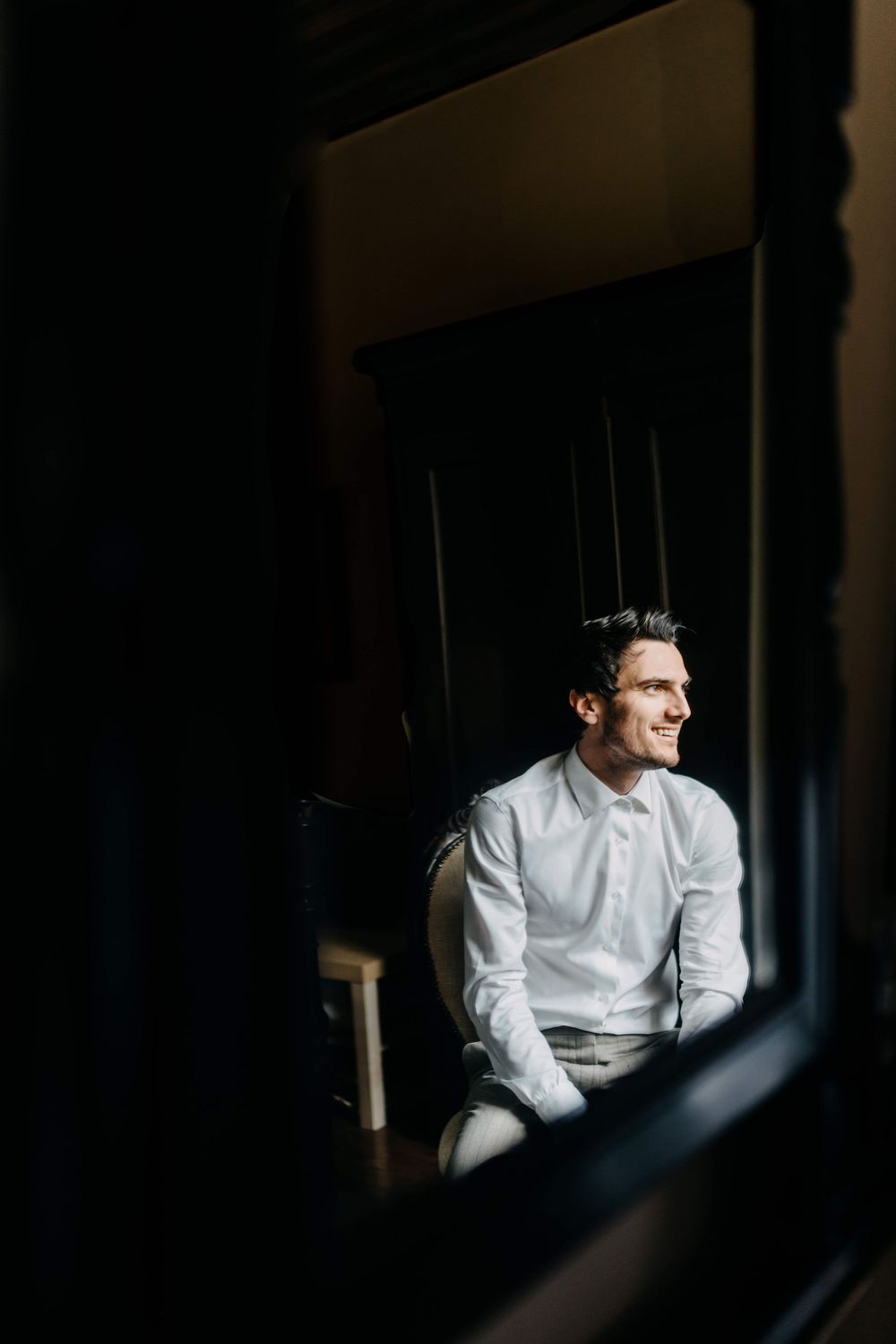 A person in a white dress shirt looks pensively through a window in dramatic lighting and shadows.