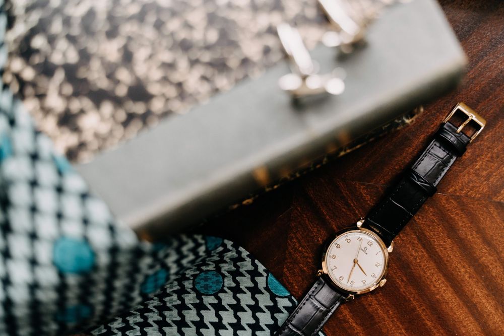 A classic wristwatch with black leather strap and golden face rests on a wooden surface next to textured fabric.