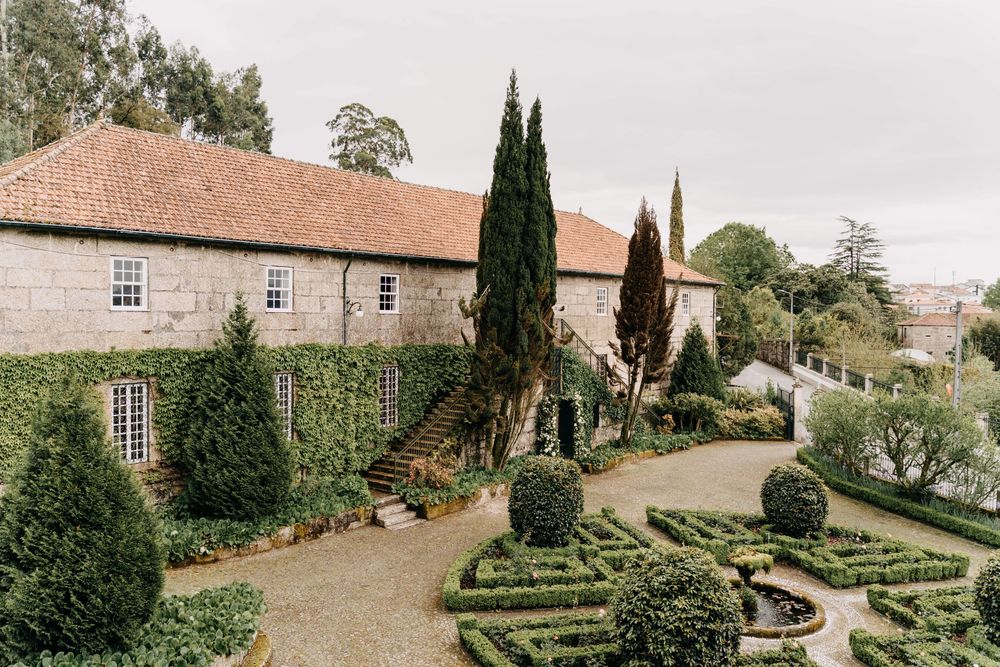 A formal garden with manicured hedges and cypress trees leads to a historic stone manor house with terracotta roof tiles.