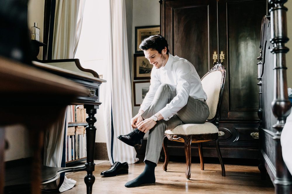 A person in formal attire puts on black dress shoes while sitting in an elegant room with a piano and white curtains.