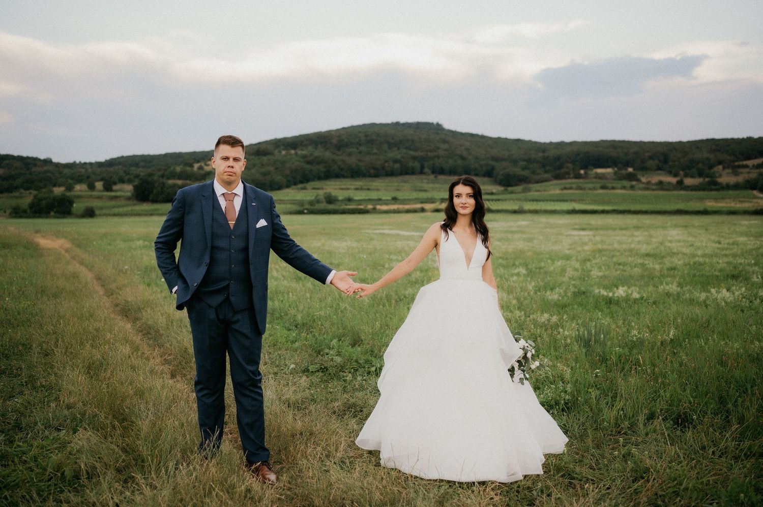 Two newlyweds stand holding hands in a beautiful grassy meadow with green hills in the background.