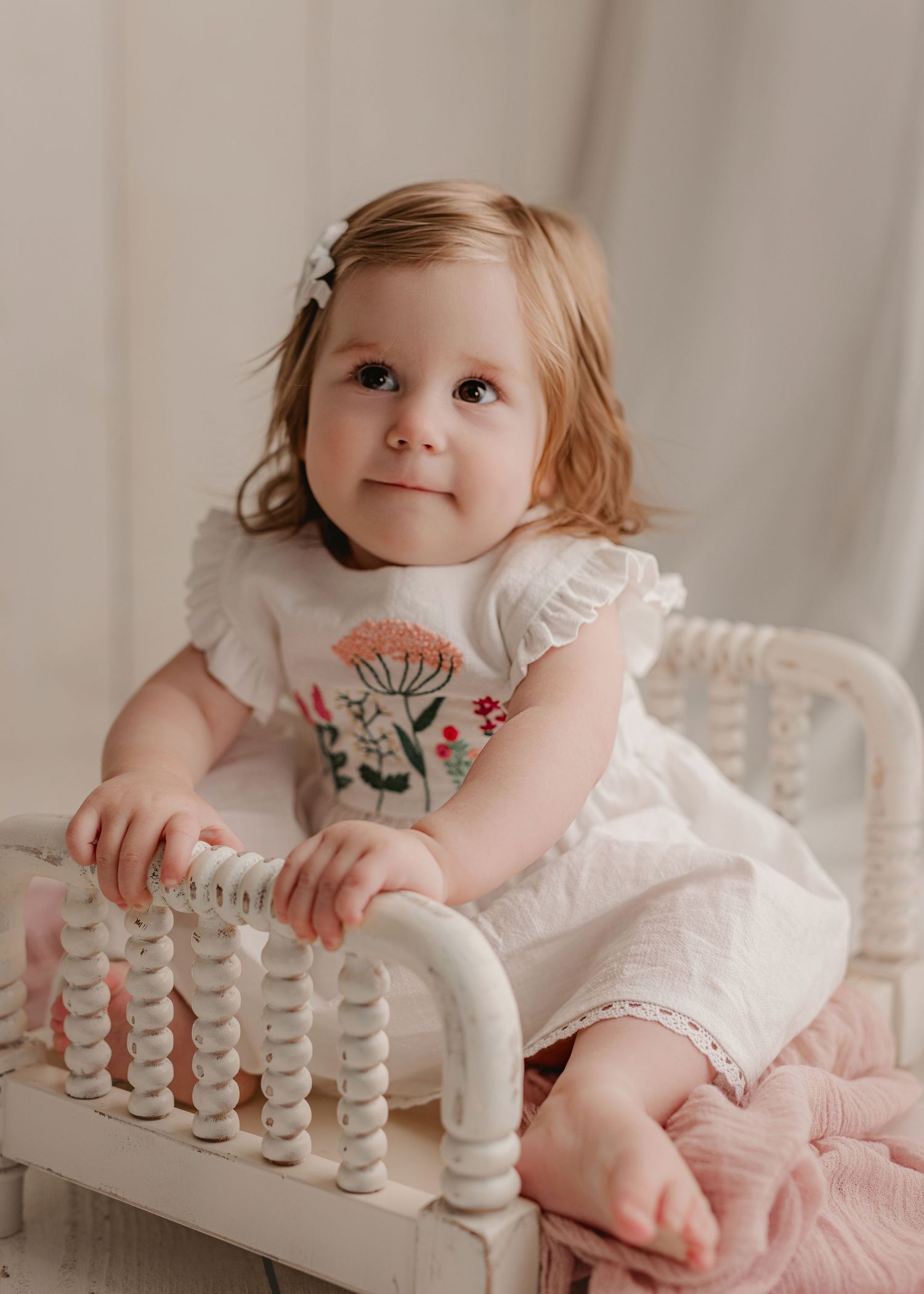 A baby in a beige knit romper sitting on a cream-colored backdrop smiling at the camera.