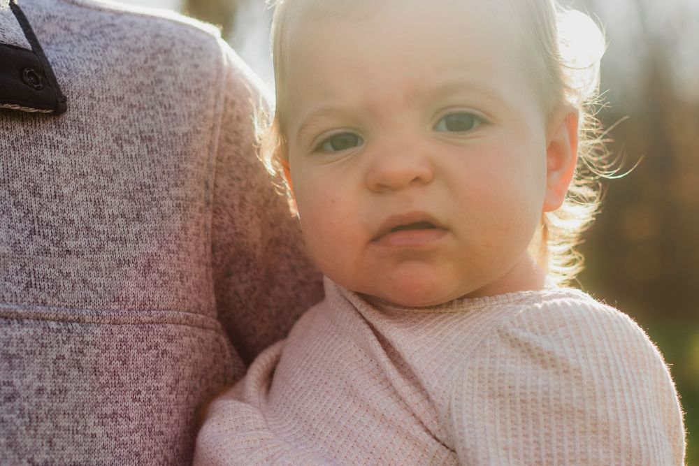 Close-up portrait of an adorable baby in a pink outfit resting on a parent's shoulder in warm sunlight.