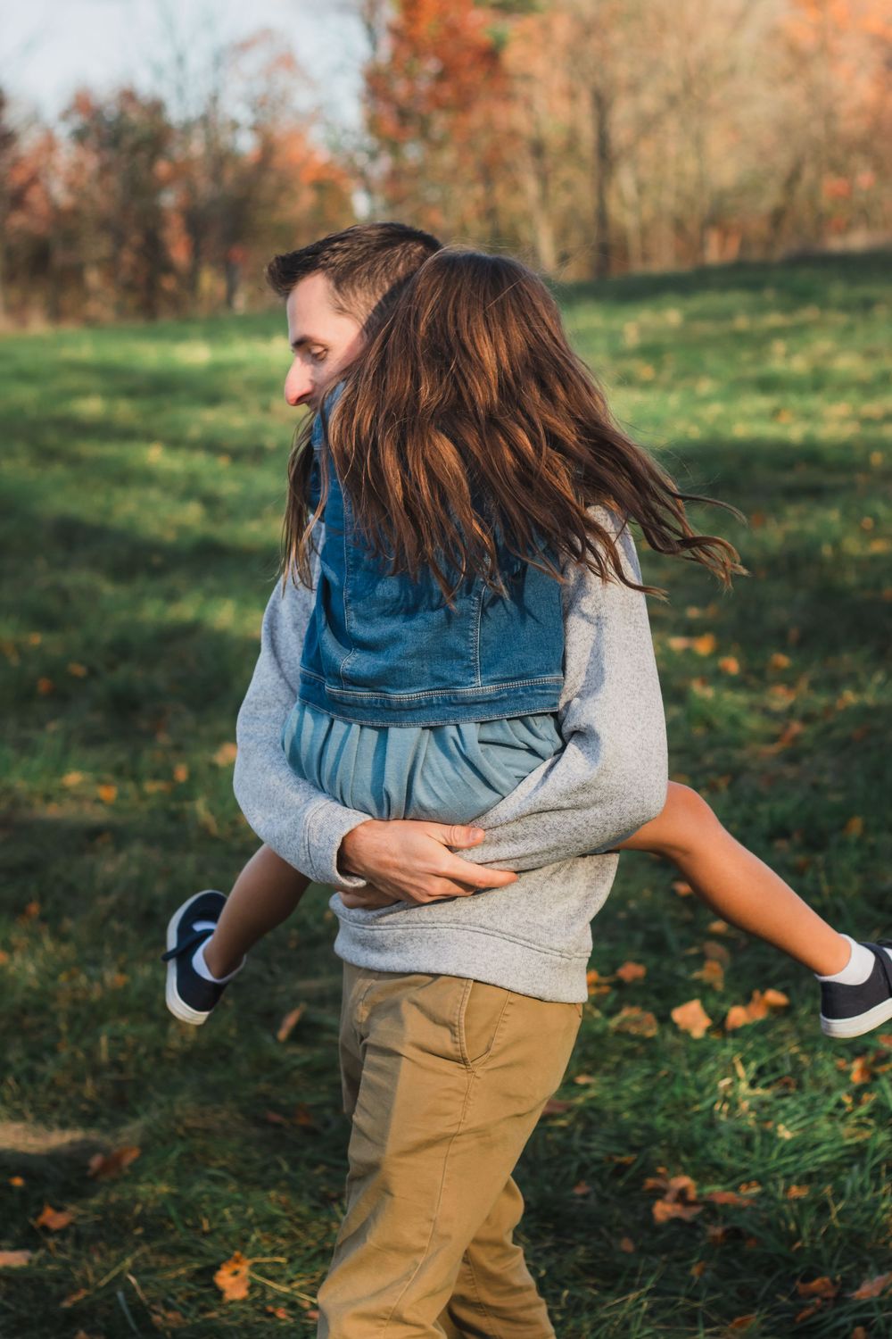 Man carrying laughing girl in denim jacket across green autumn field.