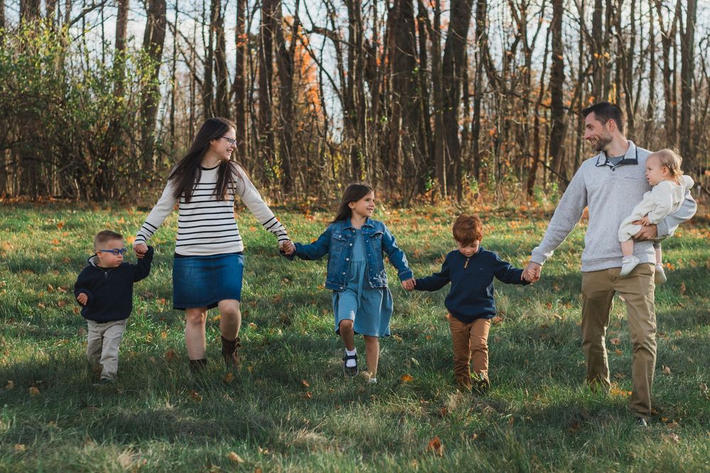 A joyful family of six holds hands and runs together through a sunny autumn park with colorful foliage.