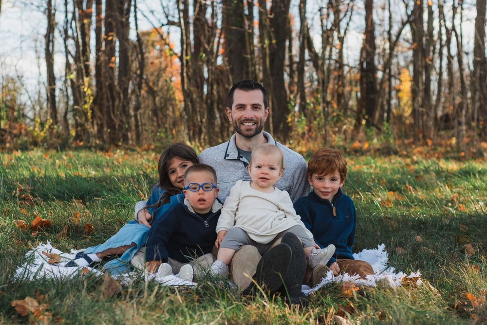 A smiling father sits on a blanket in an autumn park surrounded by his four young children.