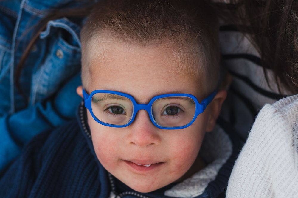 Close-up portrait of a cute baby wearing oversized blue glasses, looking up with a gentle smile.