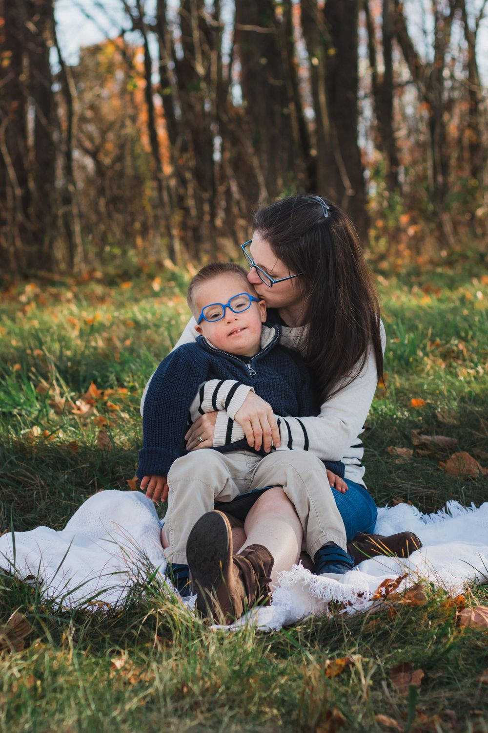 Mother kissing young boy with glasses while sitting outdoors in fall.