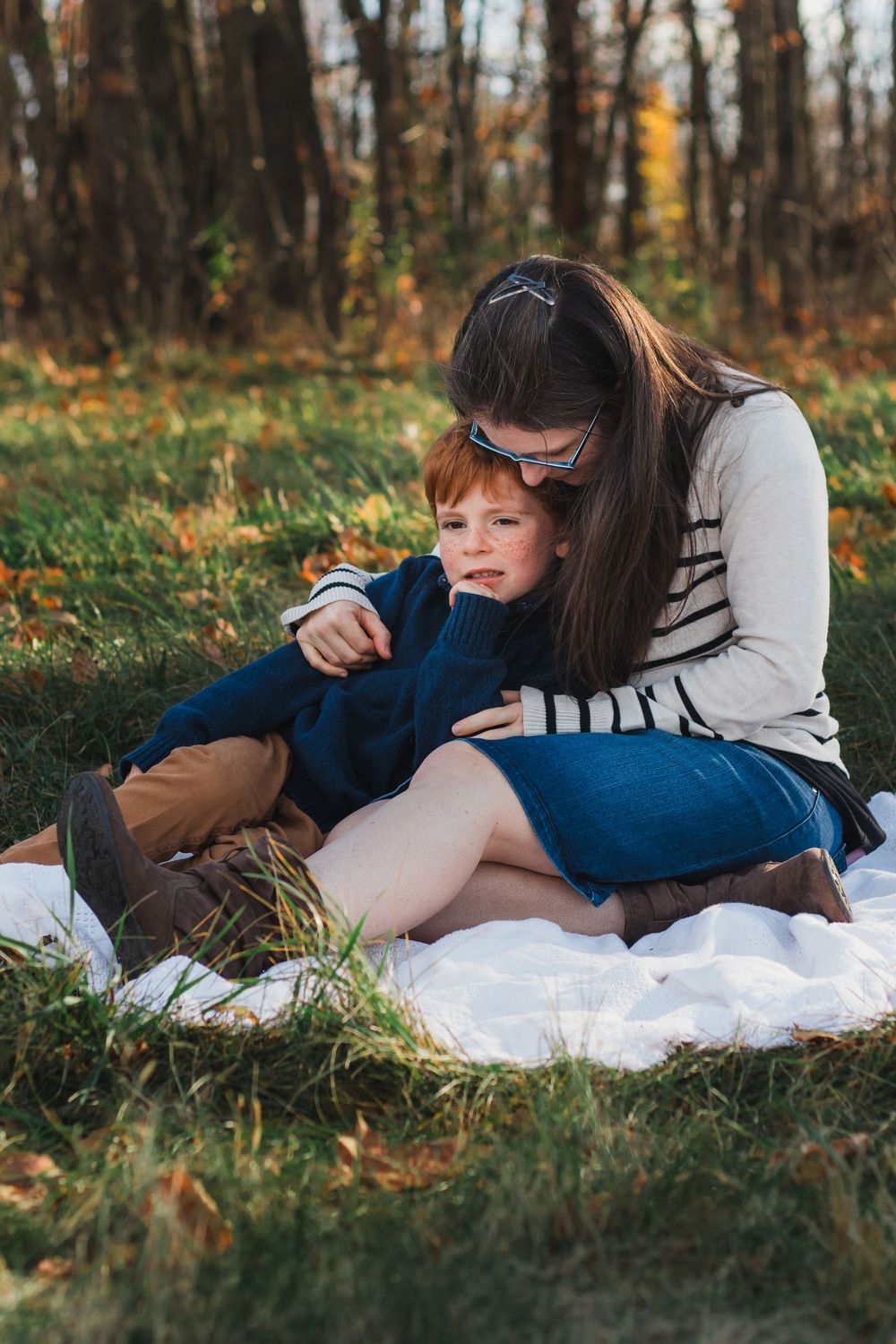 Mother embracing red-haired boy on blanket in autumn forest setting.