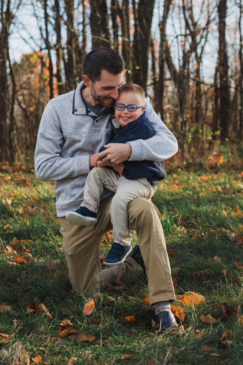 Father laughing with young boy wearing glasses in fall foliage park.