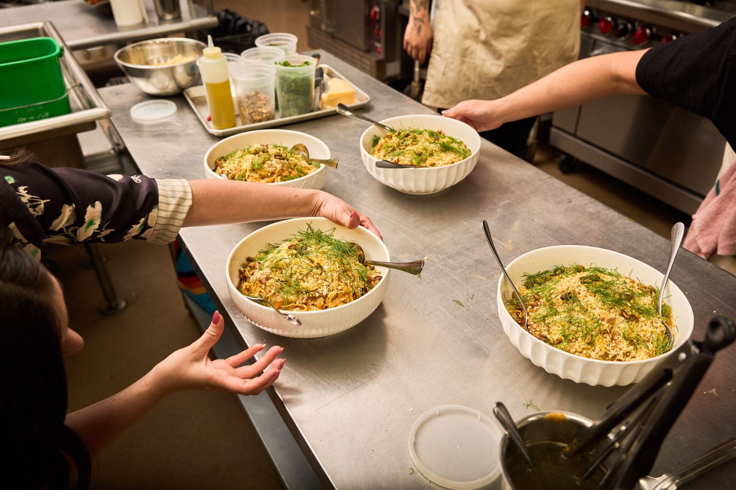 Several hands reach for portions of mac and cheese from white baking dishes on a stainless steel prep table.