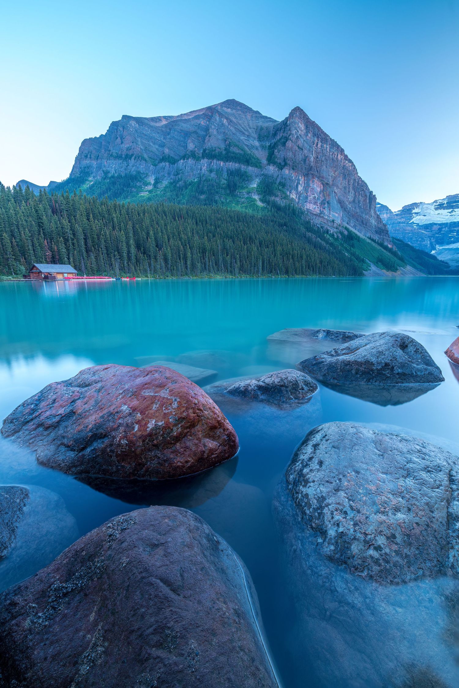 Turquoise waters of Lake Louise in Banff National Park with large boulders and mountain peaks against clear blue sky.