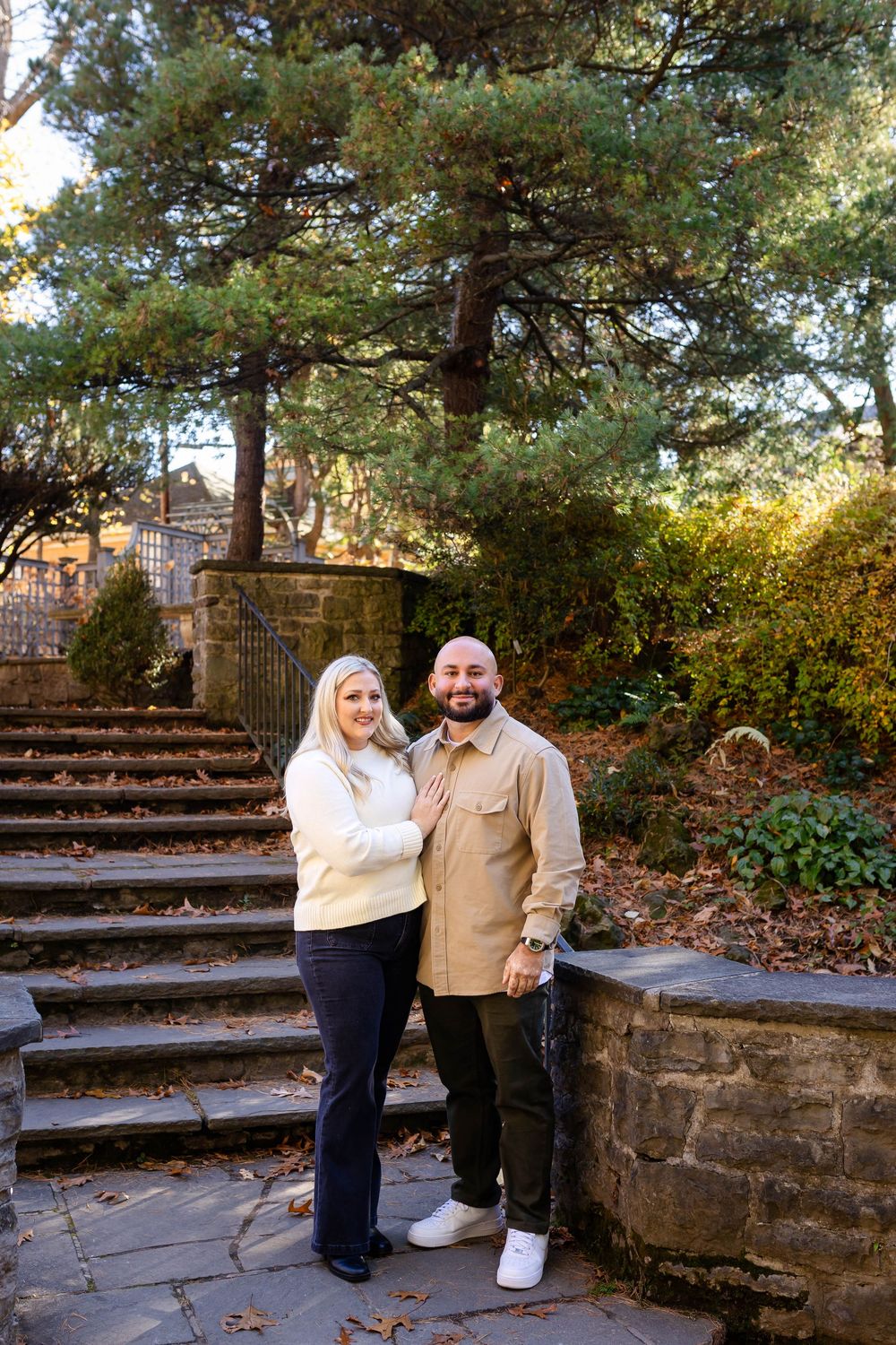 A couple stands on stone steps surrounded by autumn foliage and trees in a romantic outdoor setting.