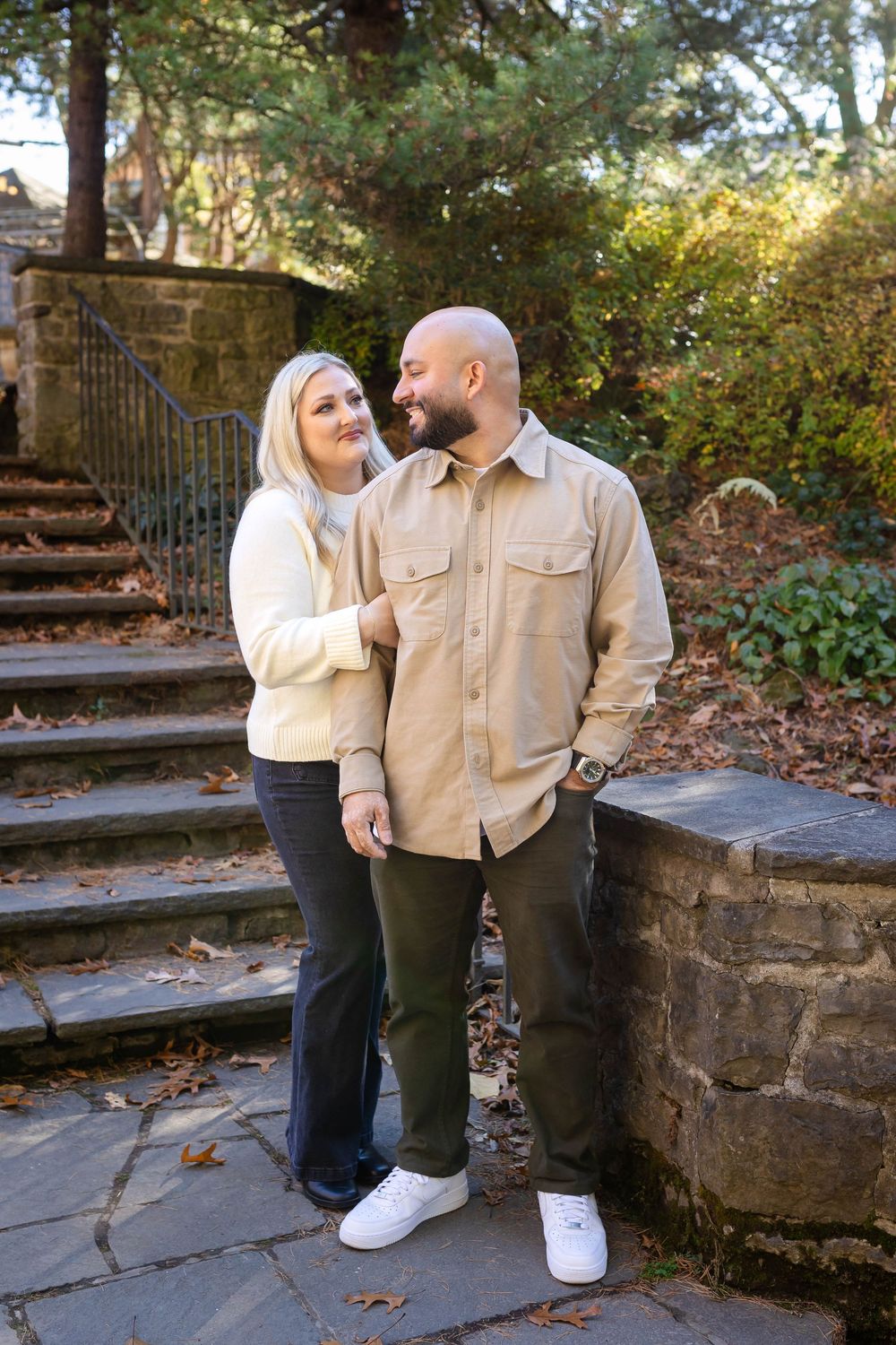 A romantic couple poses together on rustic stone steps surrounded by autumn foliage in an outdoor garden setting.
