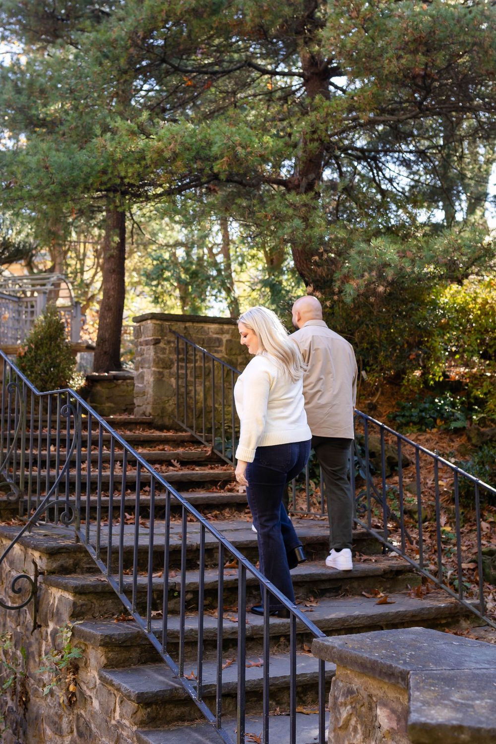 A couple walking up stone garden steps surrounded by evergreen trees and wrought iron railings on an autumn day.