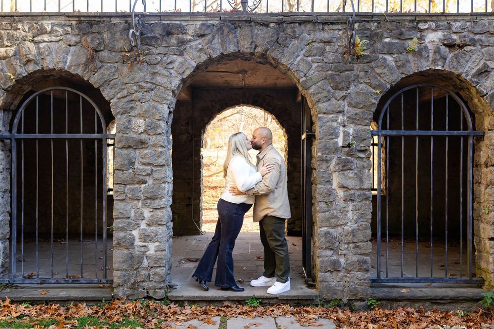 Couple embraces in stone archway between barred windows of historic building during autumn photo session.