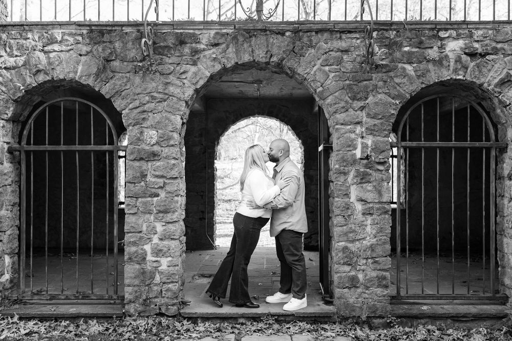 A romantic black and white photo of a couple embracing in a stone archway with iron bars.