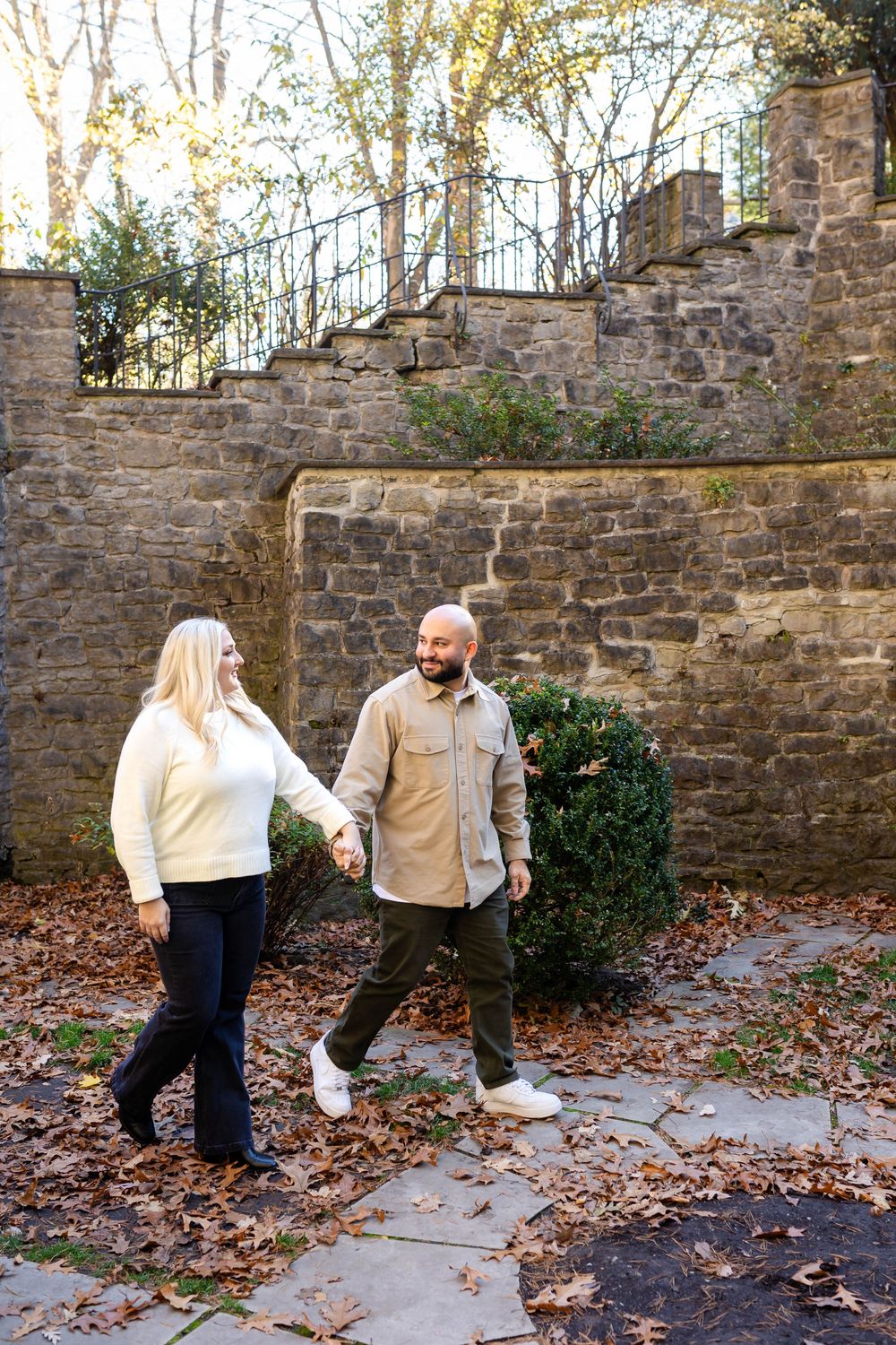 A couple strolling along a stone-walled garden path covered in fallen autumn leaves.