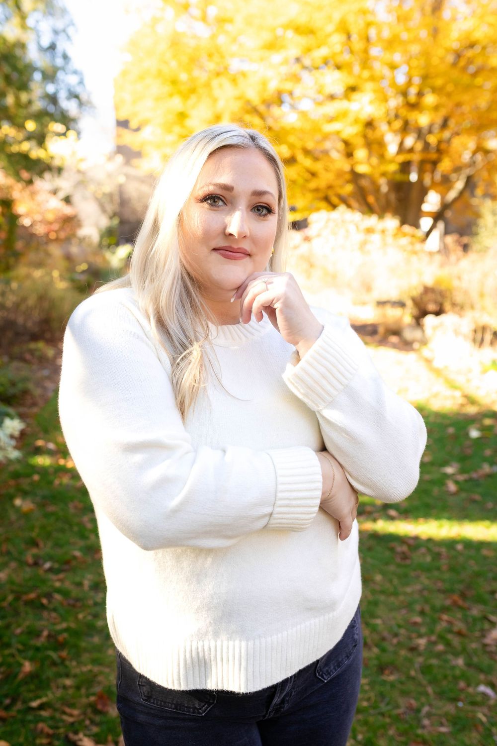 A person in a cozy white sweater poses outdoors during autumn with golden leaves in the background.