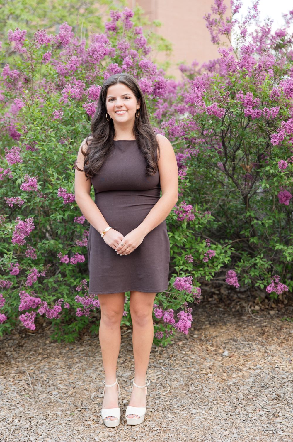 A dark gray sleeveless dress stands out against vibrant purple lilac bushes blooming in a garden setting.