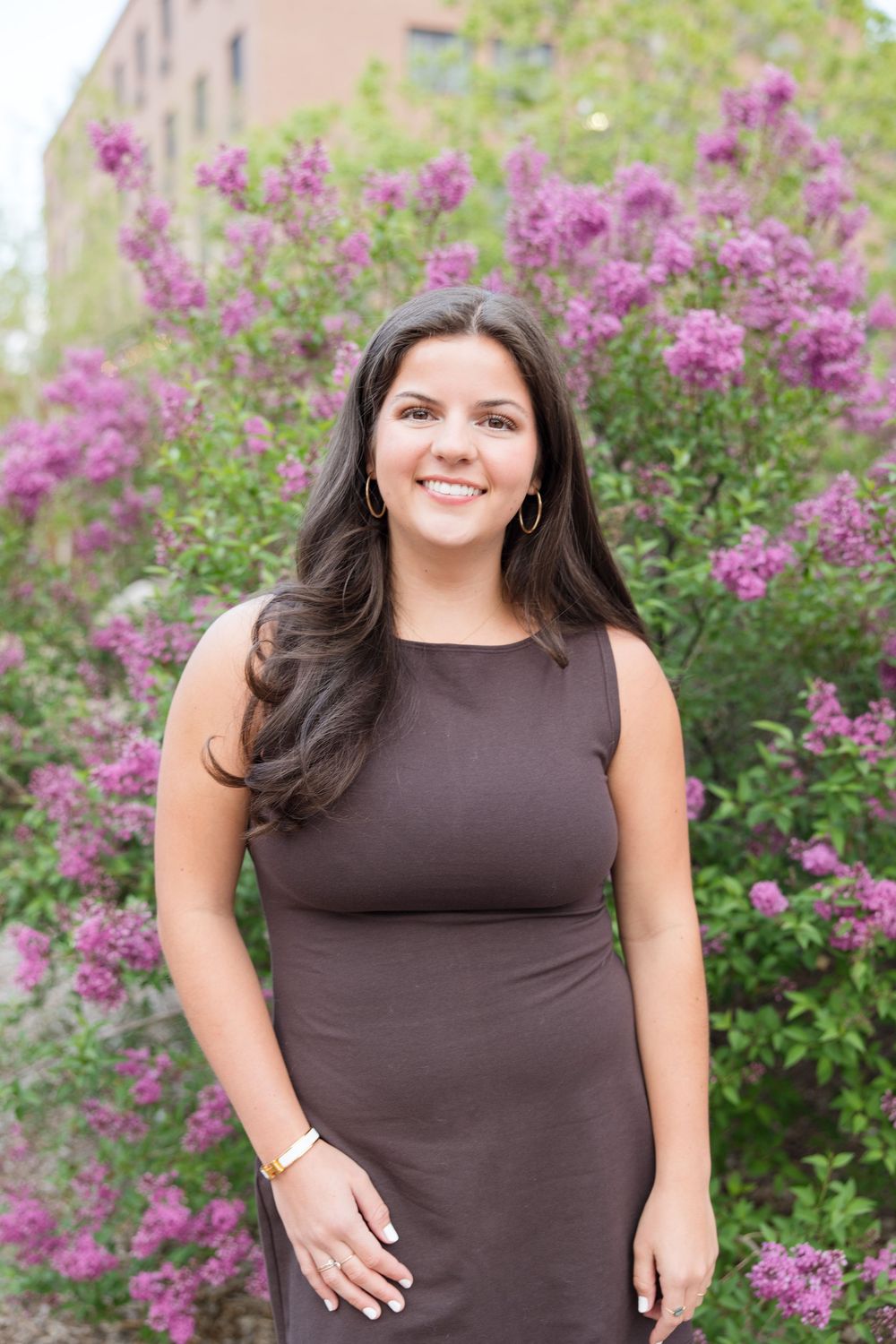 A woman in a brown sleeveless dress poses against flowering purple bushes in a garden setting.