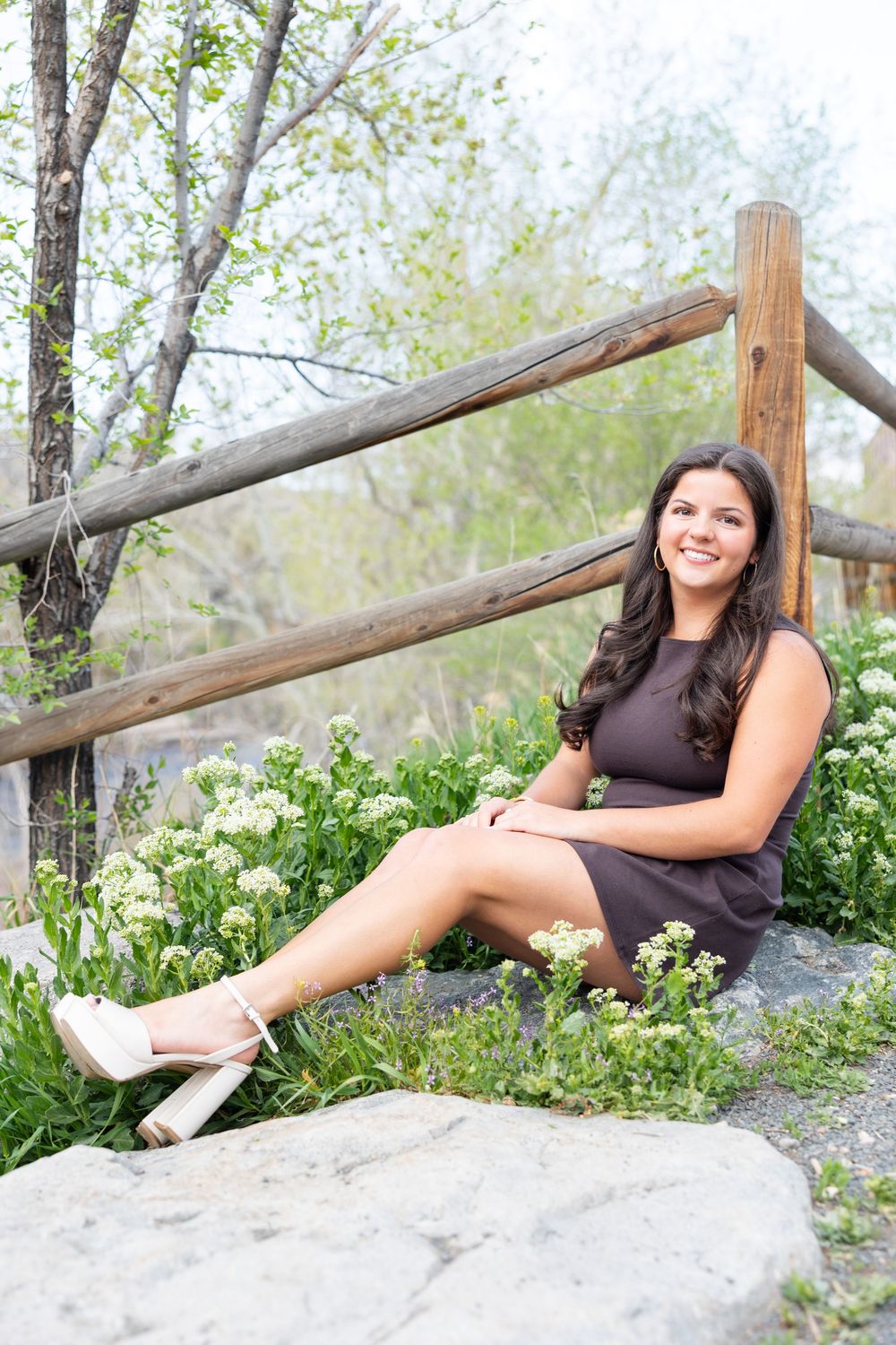 A person in a black dress sits casually on a rock by a wooden fence surrounded by spring flowers and trees.