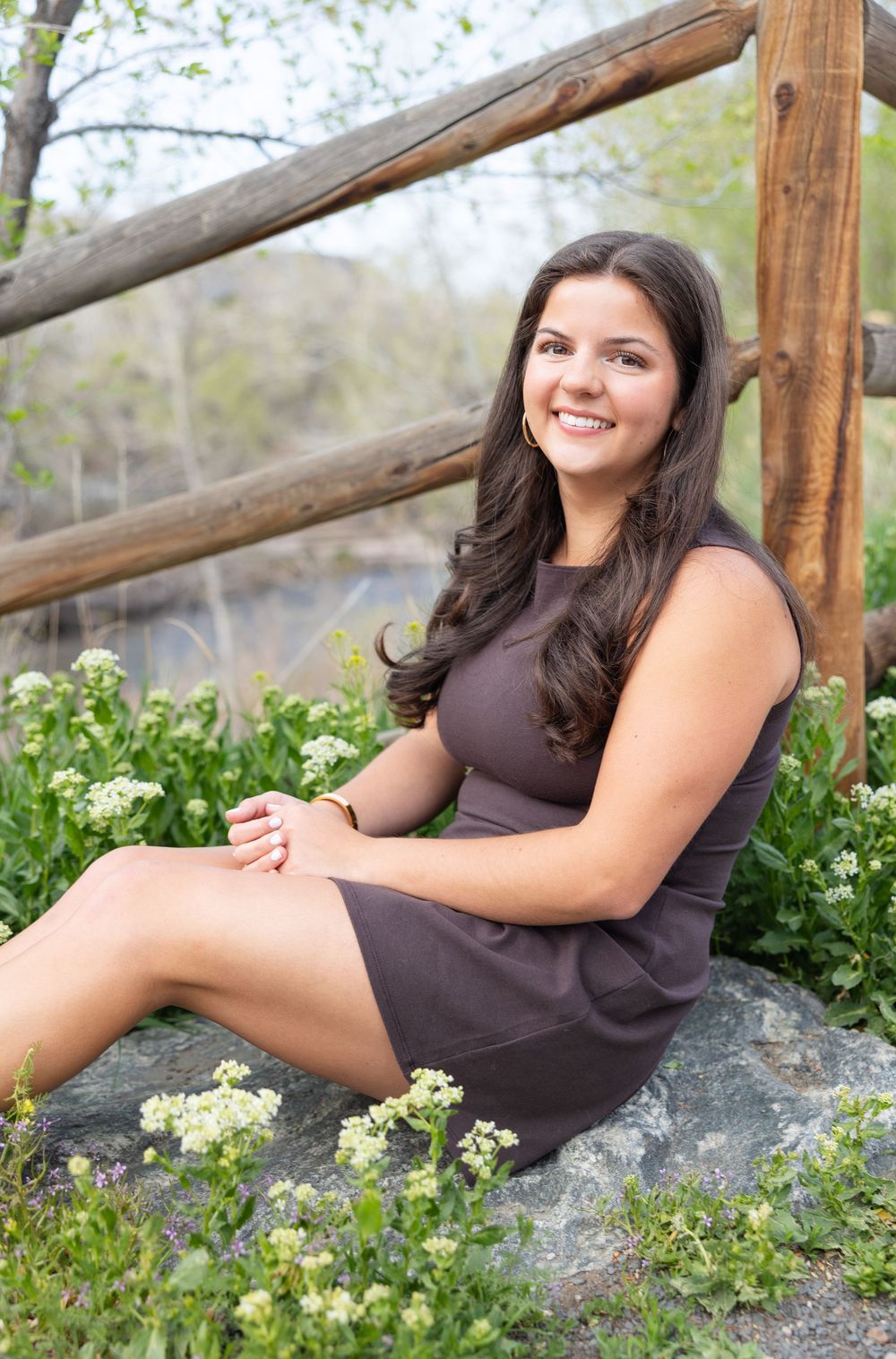 Woman in brown dress sitting on rock surrounded by white wildflowers and wooden fence in outdoor setting.