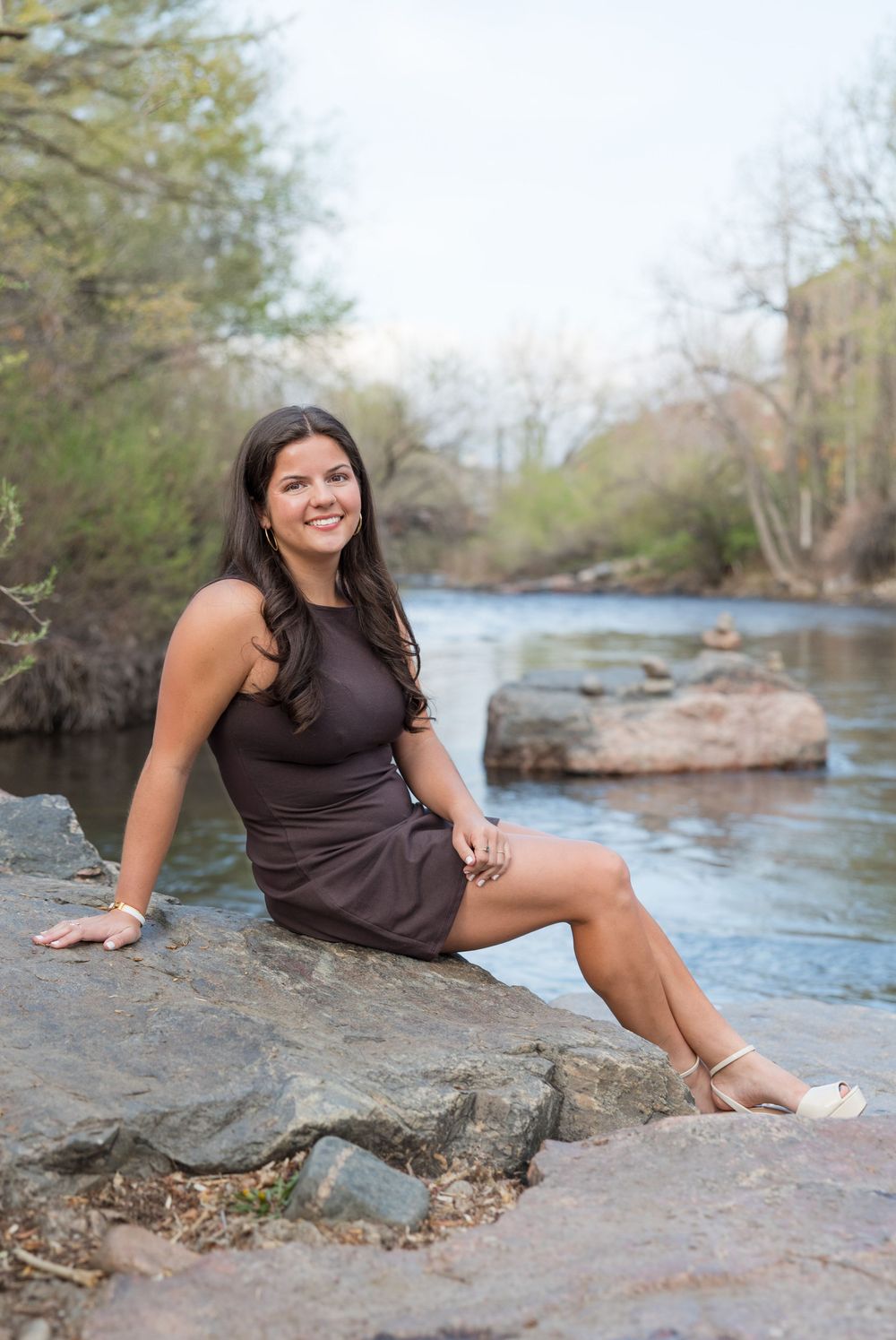 A person in a black dress sits barefoot on rocky outcrop along a scenic river on a spring day.