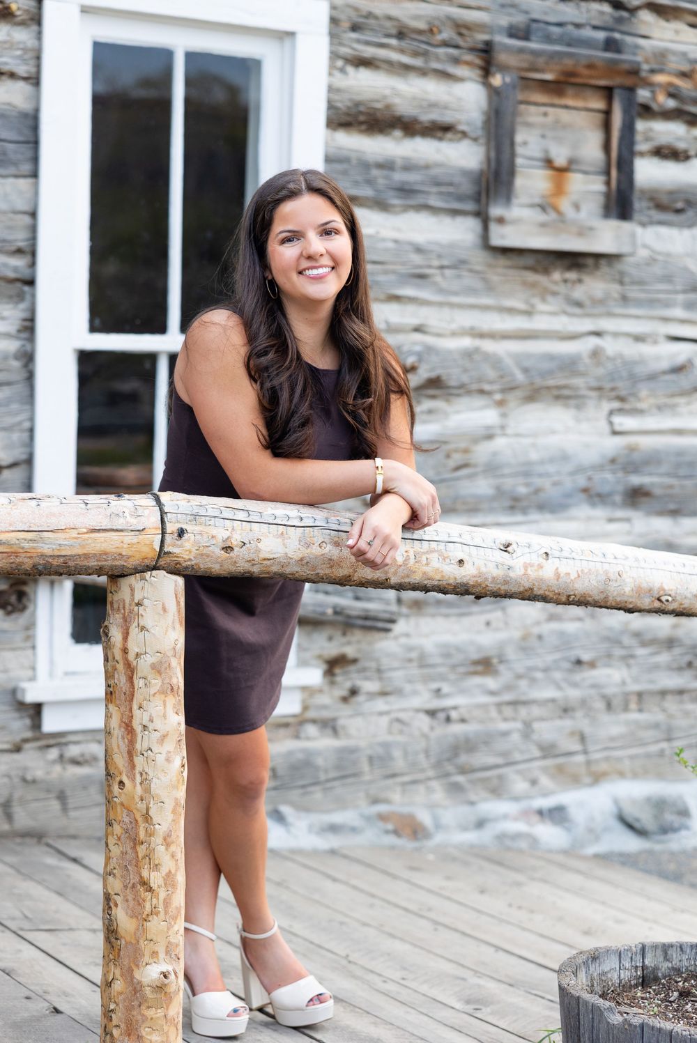 A person in a black dress leans on a rustic wooden railing outside a log cabin with weathered wood walls.