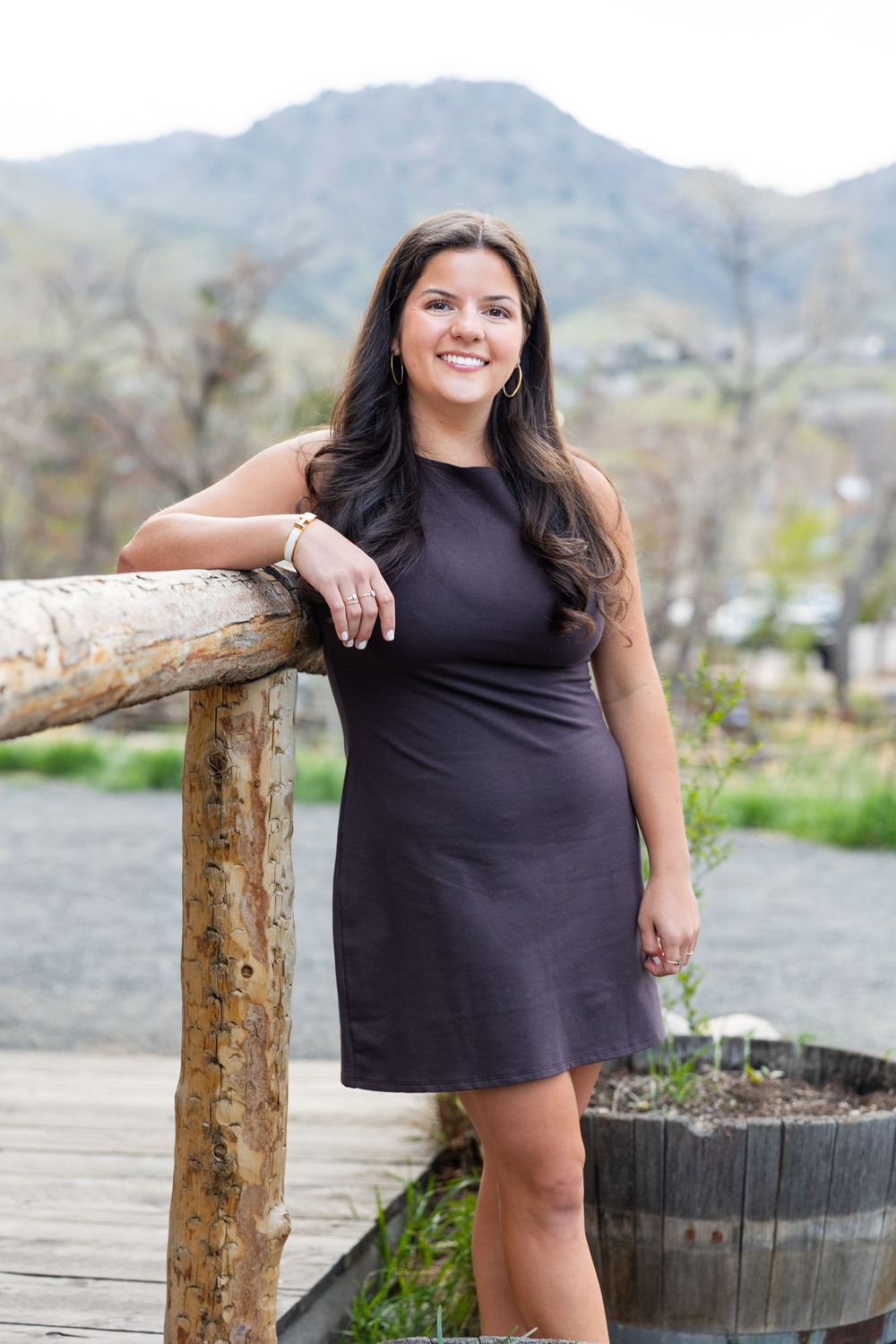 A woman in a dark dress leans against a rustic wooden railing with mountains in the background.