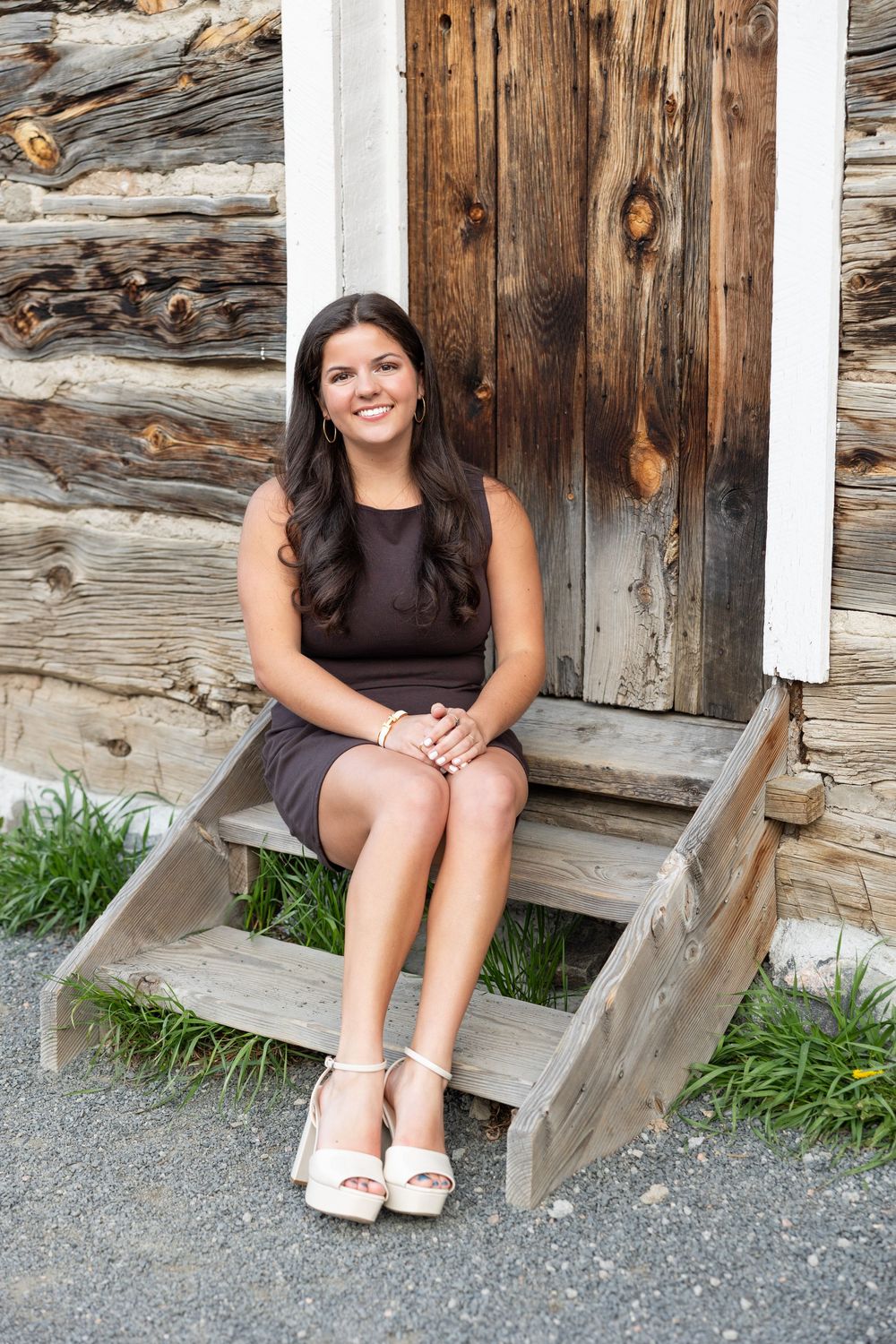A woman in a dark dress sits on wooden steps outside a rustic log cabin.