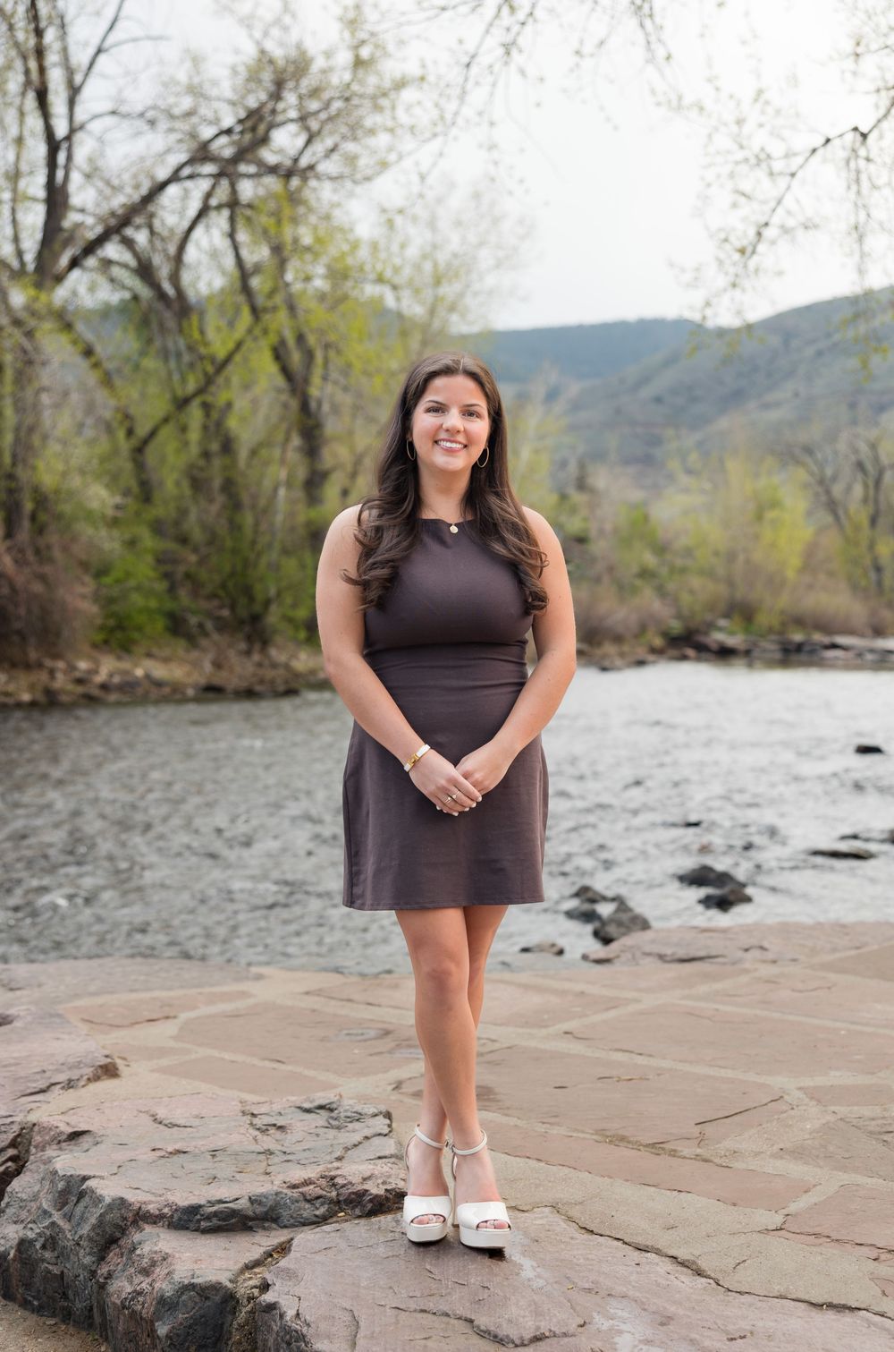 Someone in a purple dress stands on rocky terrain beside a river with mountains and trees in the background.