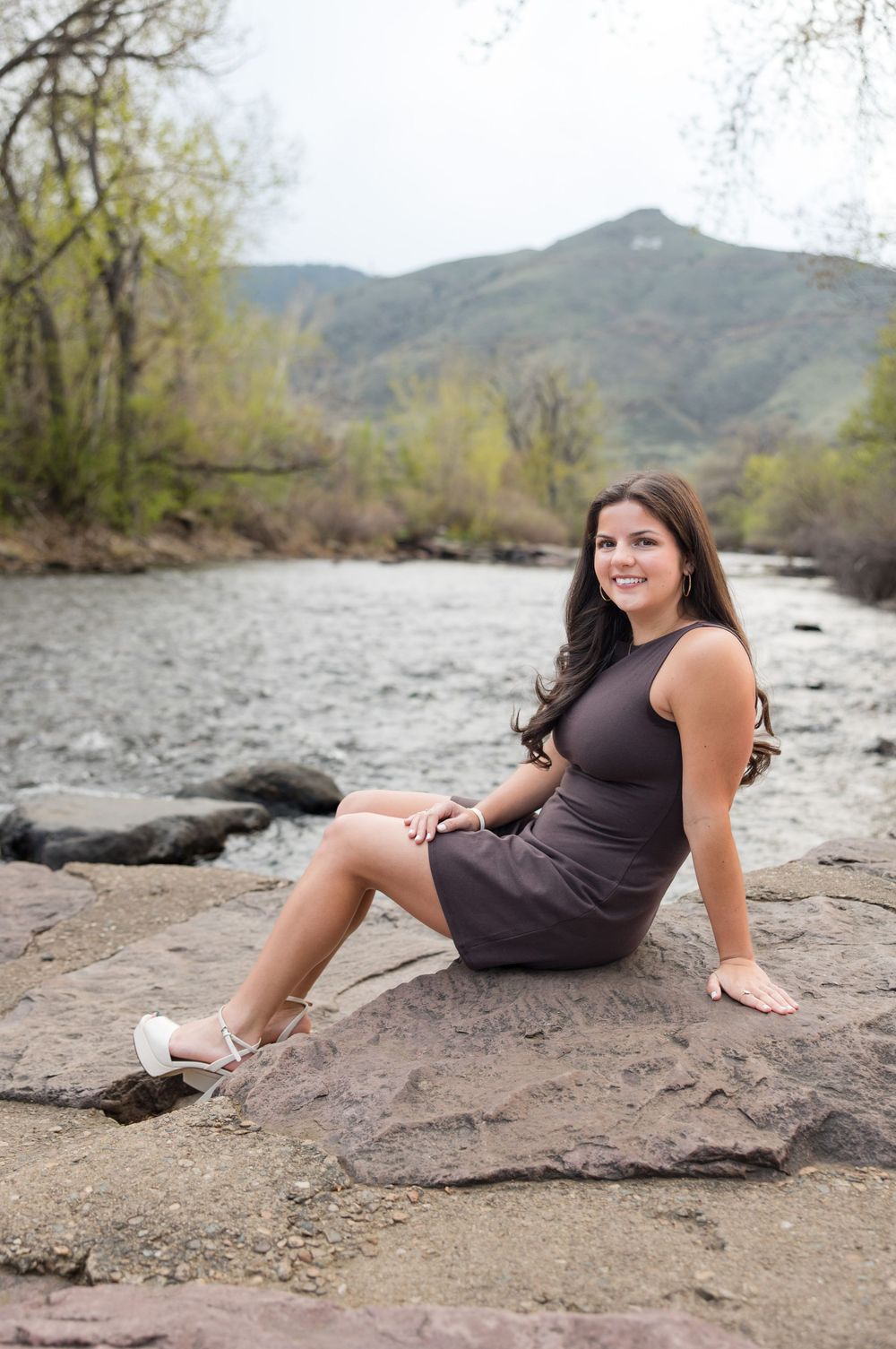 A young woman in a purple dress sits on rocks by a mountain river on a spring day.