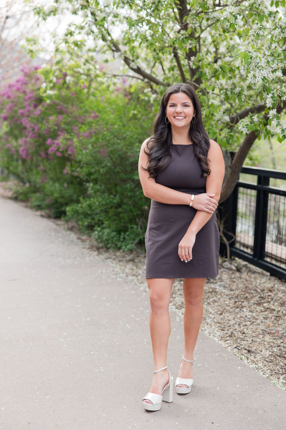 A figure in a dark sleeveless dress and platform sandals poses on a path with blooming spring trees in the background.
