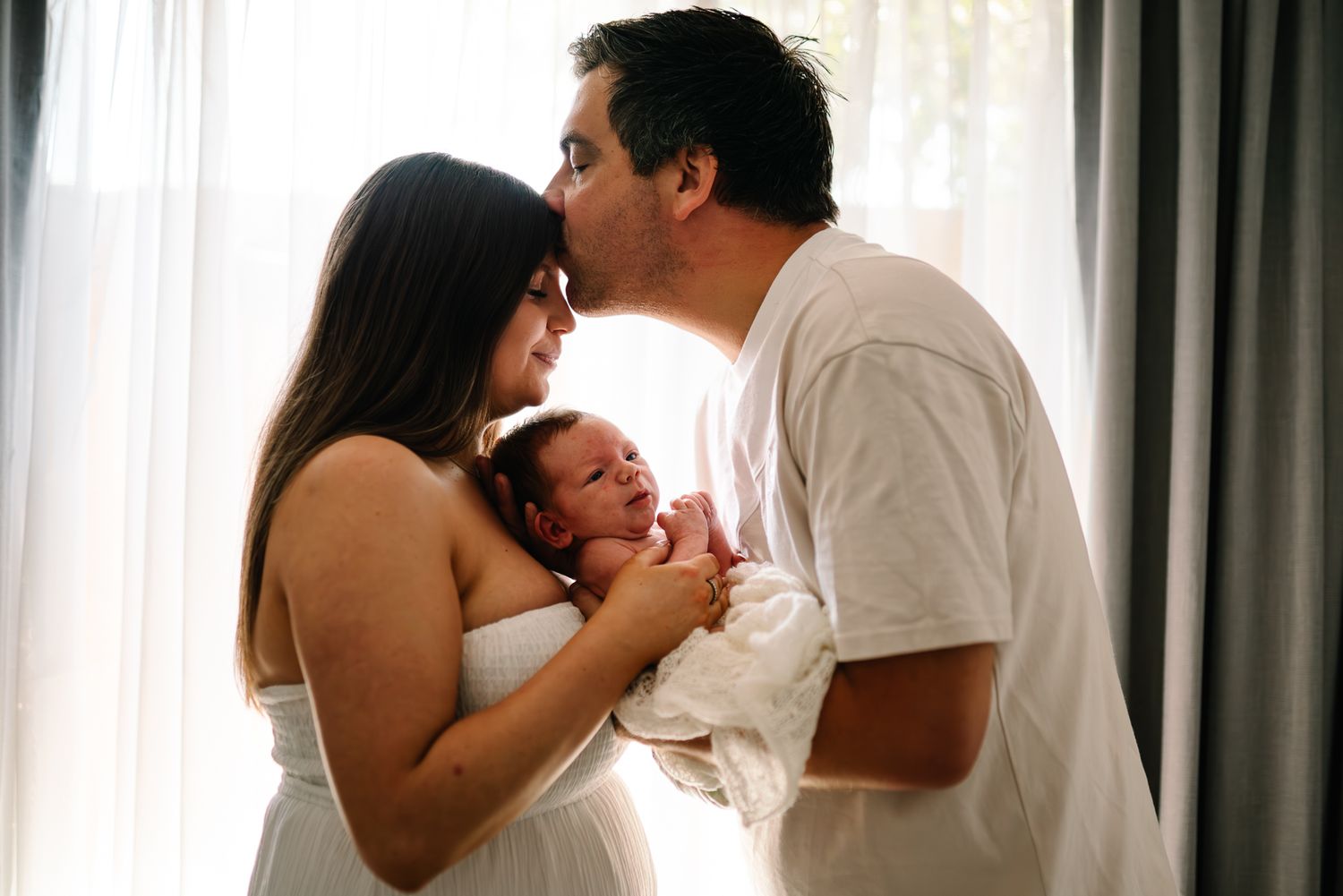 A couple embraces their newborn baby together near a sunlit window with sheer curtains taken by Melbourne newborn photographer.