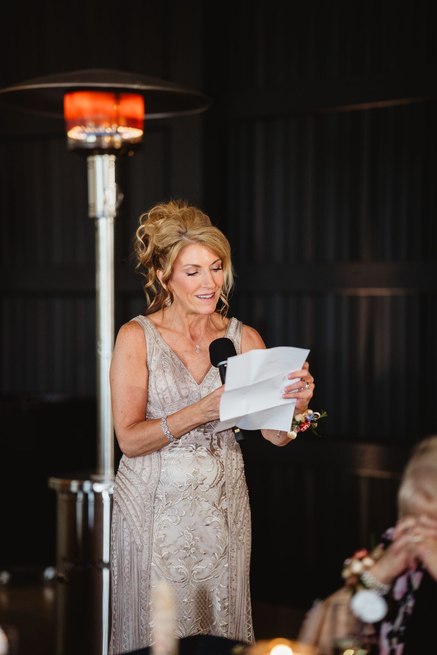 A person in a shimmering beige dress reads from papers while speaking into a microphone at an evening event.