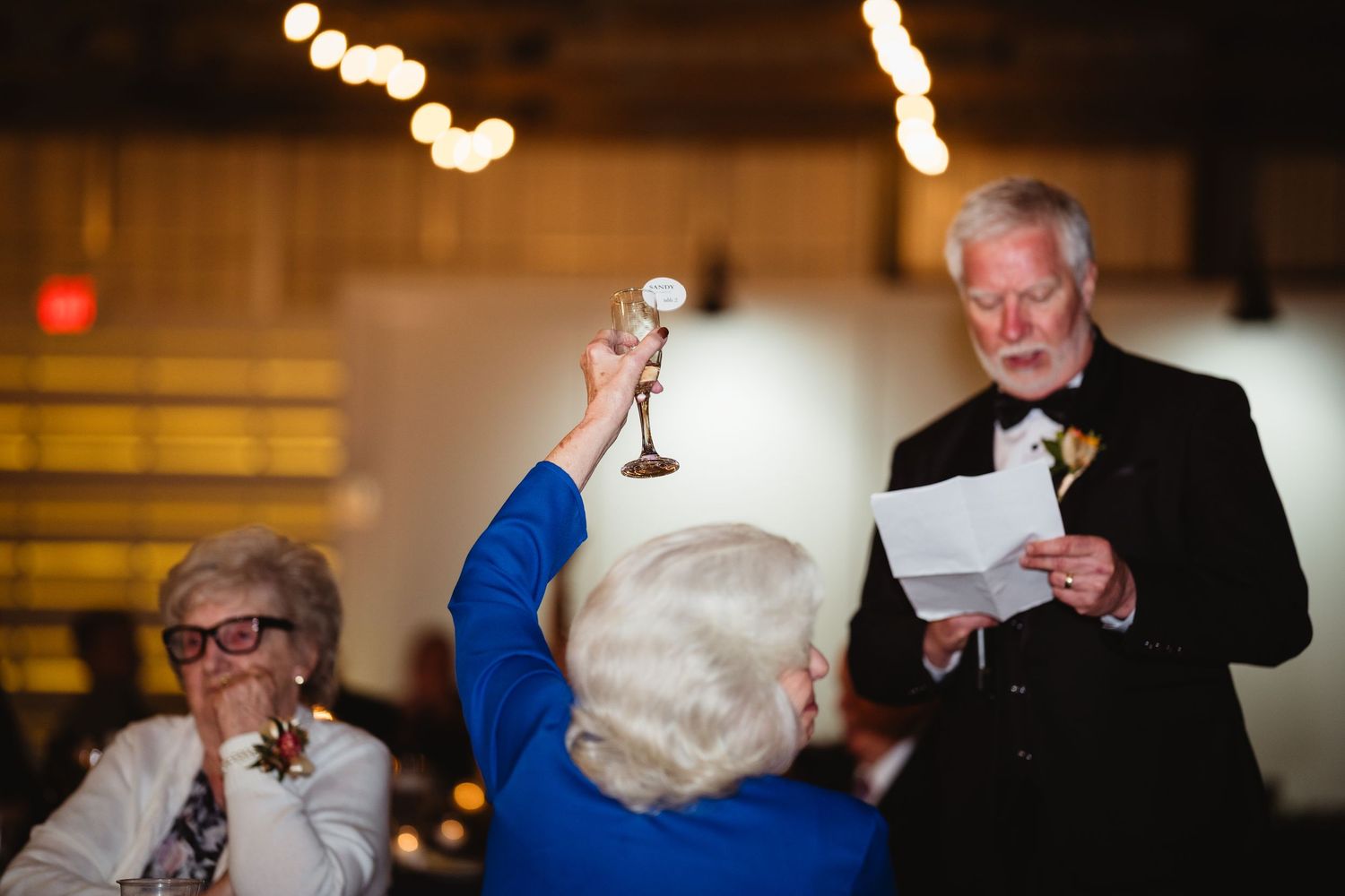 People giving toasts and reading speeches at an evening celebration event with blurred lights in the background.