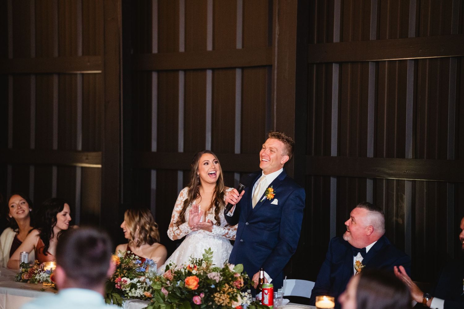 Wedding reception guests share a laugh at a rustic barn venue with warm lighting and floral table decorations.