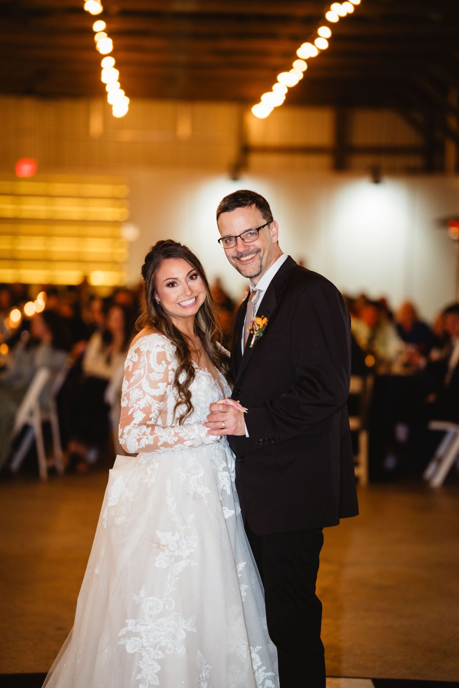 Couple poses together during wedding reception with warm string lights glowing overhead in venue.