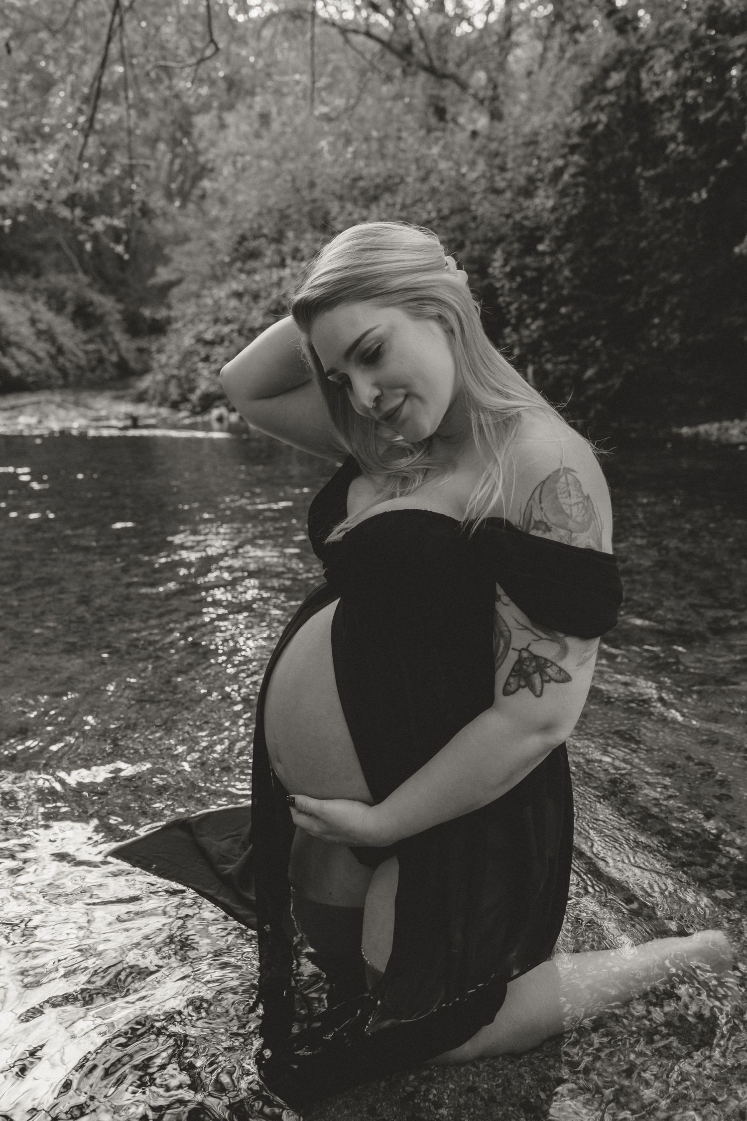 A pregnant woman in a black dress poses in a creek surrounded by lush greenery during a maternity photoshoot.